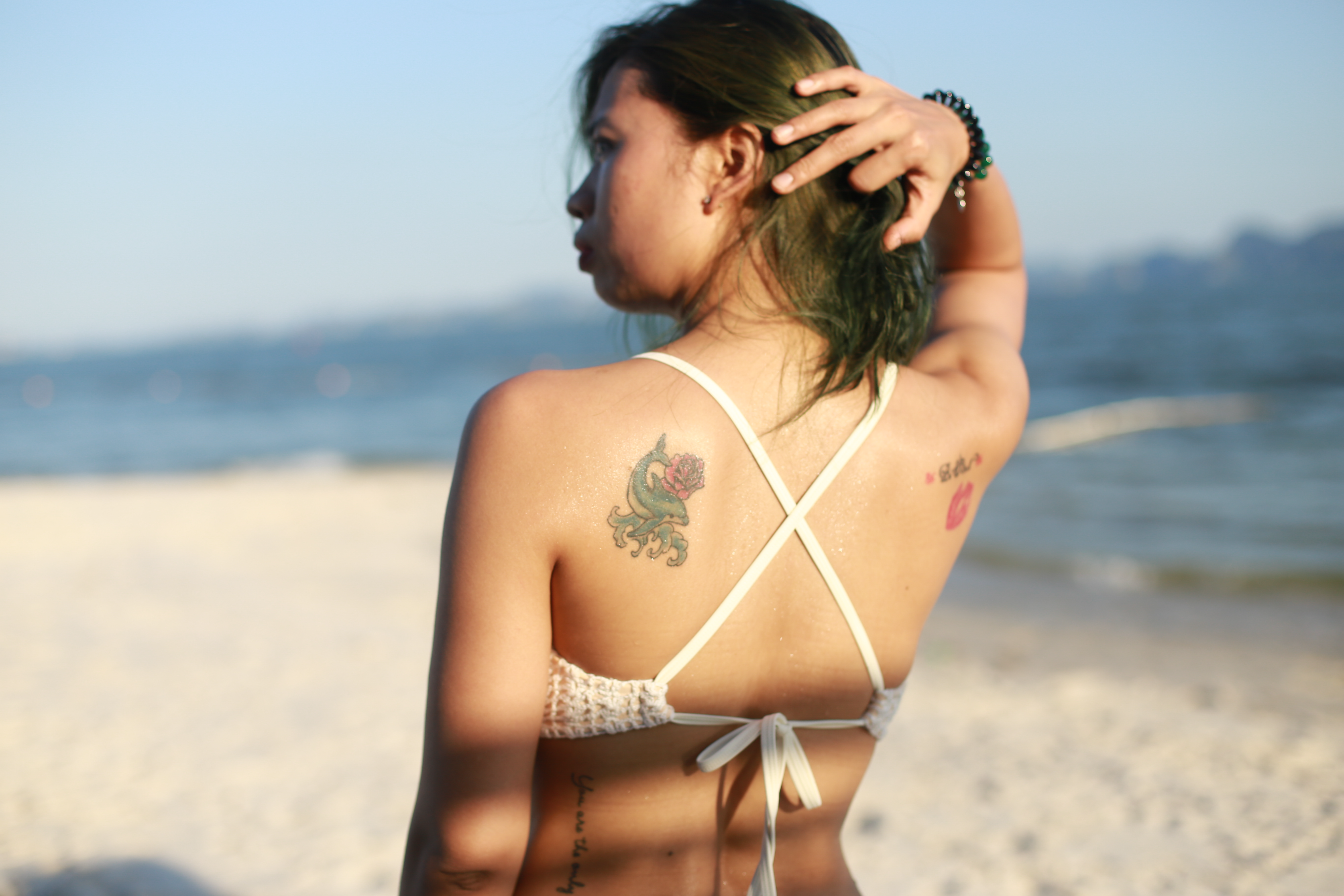 A woman in swimwear with tattoos relaxes on a sunny beach looking out at the ocean.