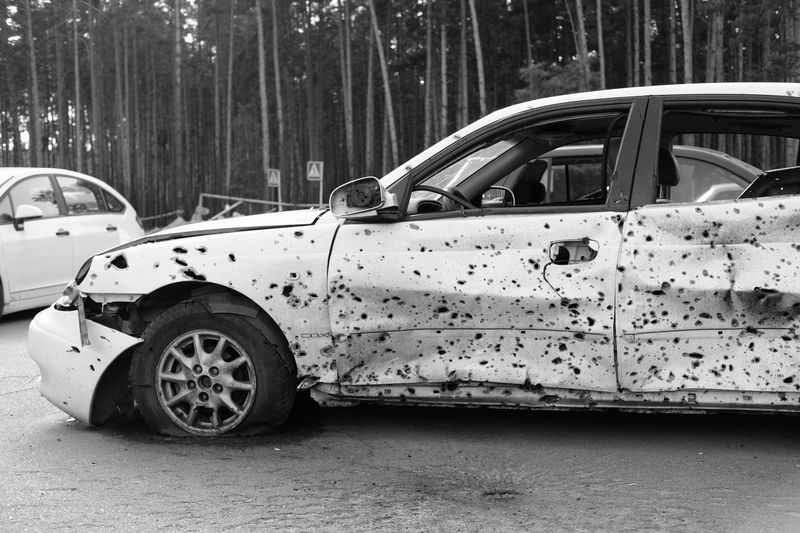 A monochrome image of a bullet-ridden car in a forest, conveying decay and abandonment.
