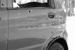 Close-up of a car door with bullet holes and a flat tire in a forest setting.