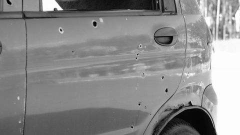 Close-up of a car door with bullet holes and a flat tire in a forest setting.