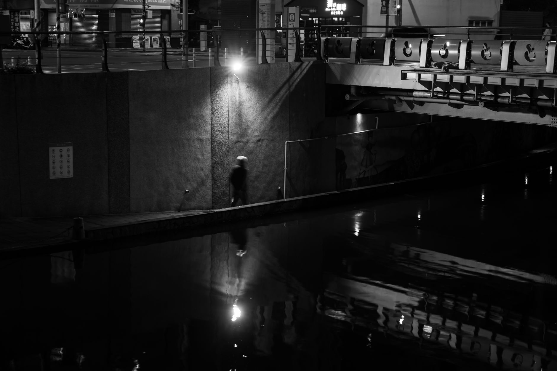 Moody urban night scene of a lone walker by a quiet waterfront, captured in black and white.