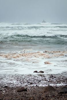 Large ocean waves crashing on a rocky shore with a distant lighthouse under a moody sky.