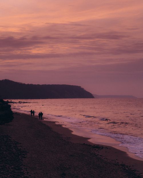 Free Silhouettes stroll along a tranquil beach at twilight, capturing the serene beauty of waves and sky. Stock Photo