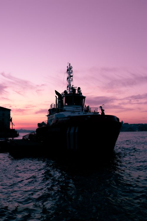 Free Tugboat silhouetted against a vibrant sunset in the harbor, creating a dramatic scene. Stock Photo