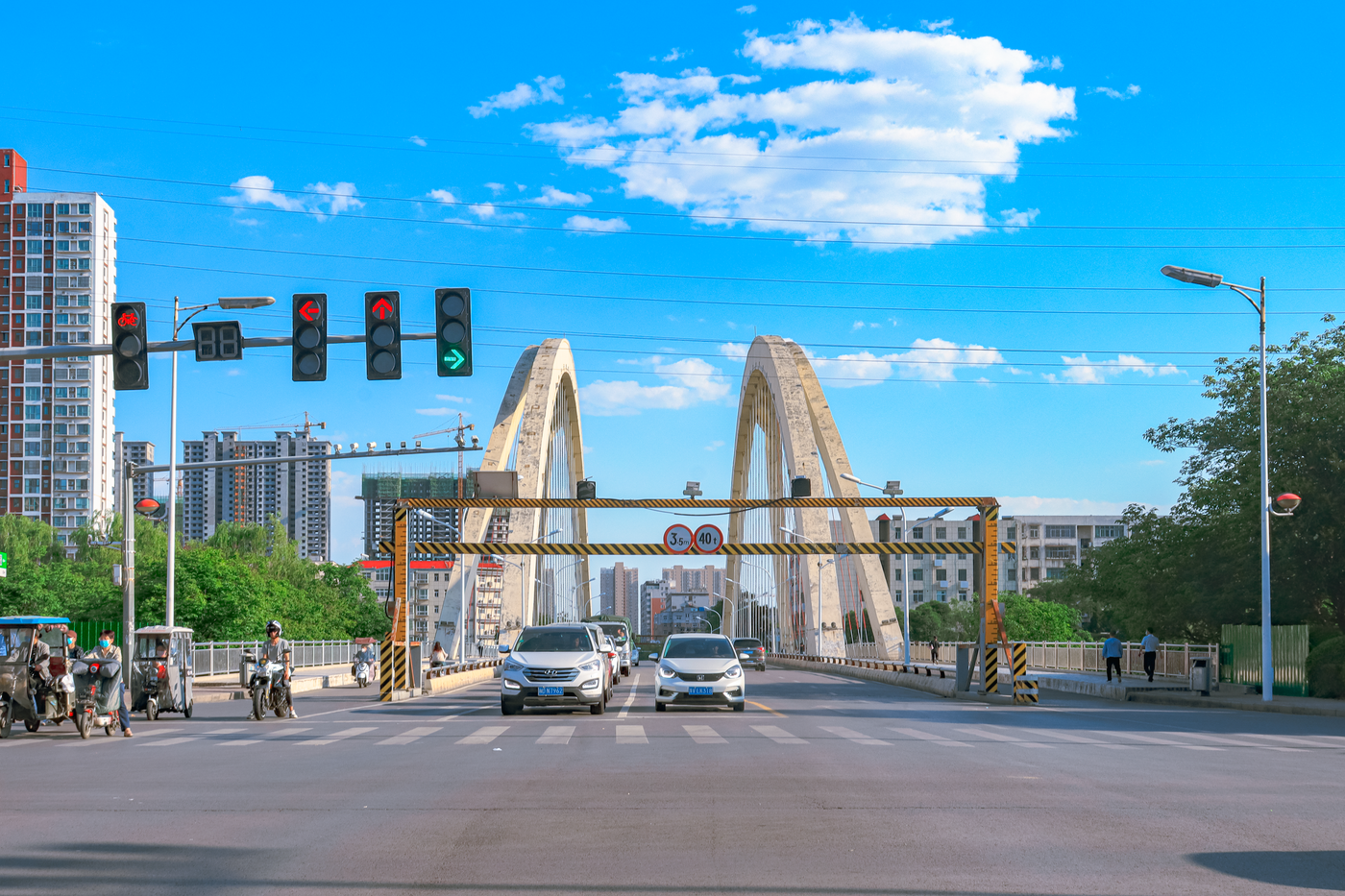 Street view of a busy urban bridge in Pingdingshan, Henan, China with vehicles and pedestrians under a clear blue sky.