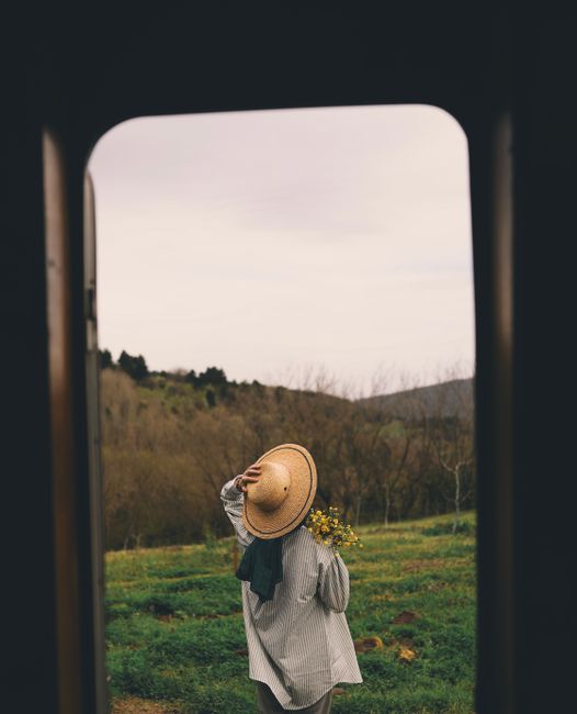 A woman wearing a wide-brimmed hat holds flowers while standing in a lush meadow viewed through a window frame.