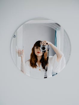 A woman capturing her reflection in a mirror while holding a glass in one hand and a camera in the other.