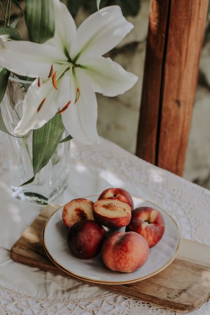 Plate of fresh peaches next to a white lily on lace tablecloth in a calm, natural light setting.