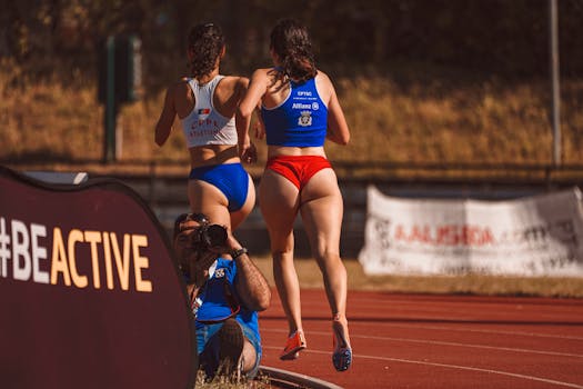 Two female athletes running on a track during a sports competition, captured by a photographer.