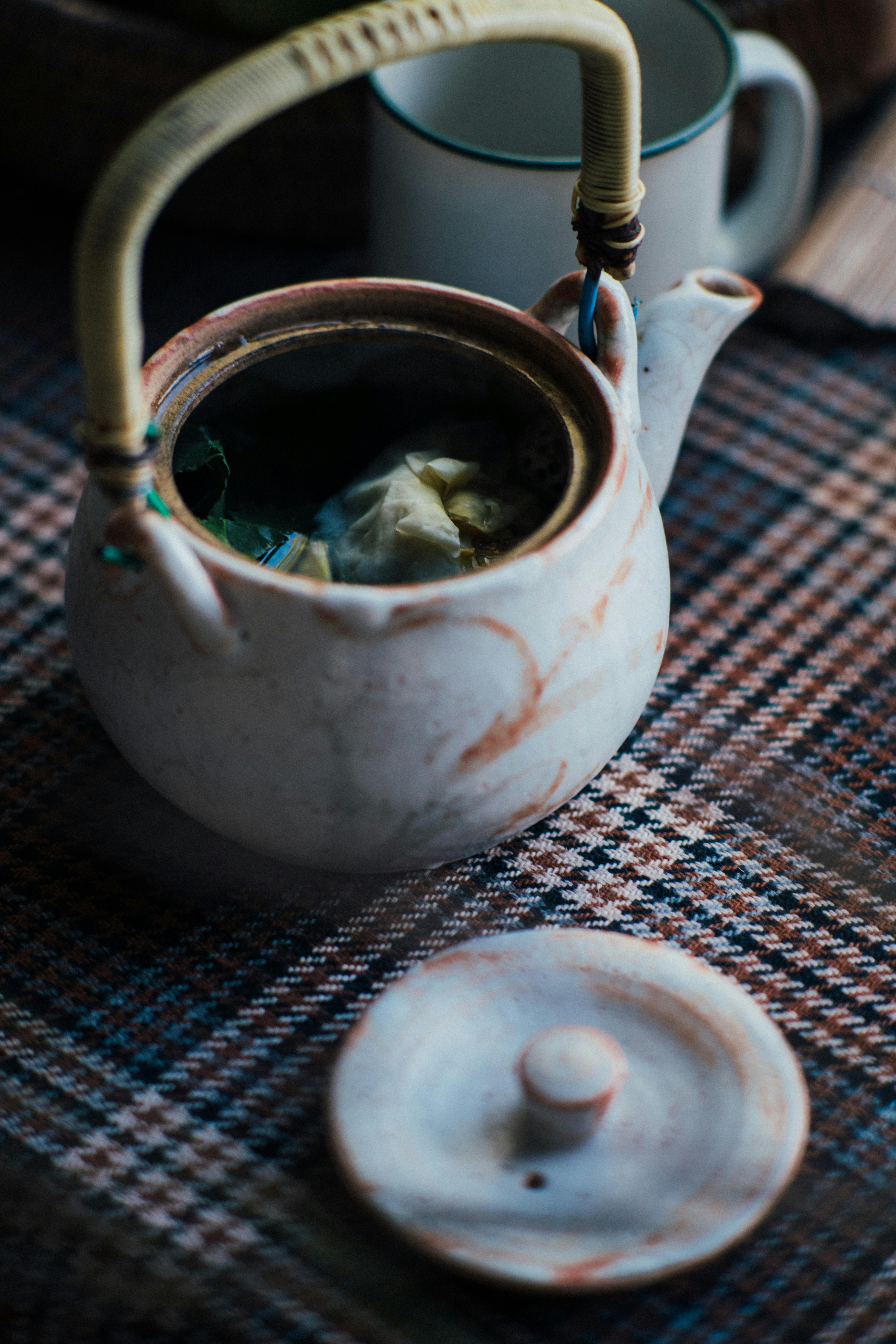 A close-up view of a ceramic teapot with an open lid on a patterned mat indoors.
