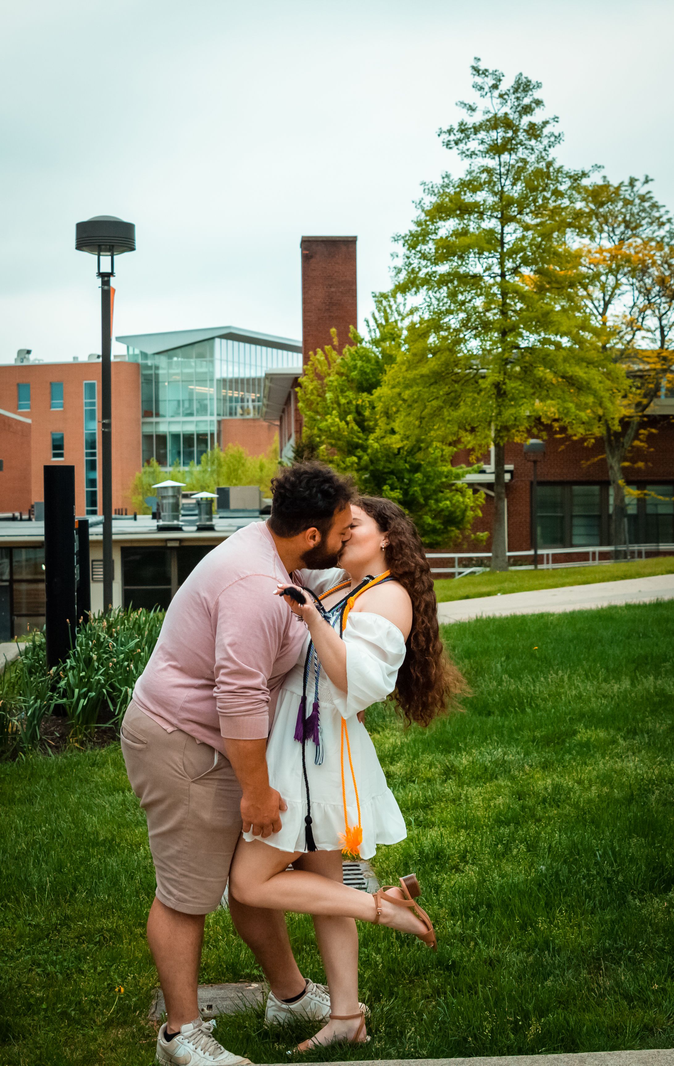 An affectionate couple shares a kiss outdoors on a lush college campus, surrounded by modern buildings.