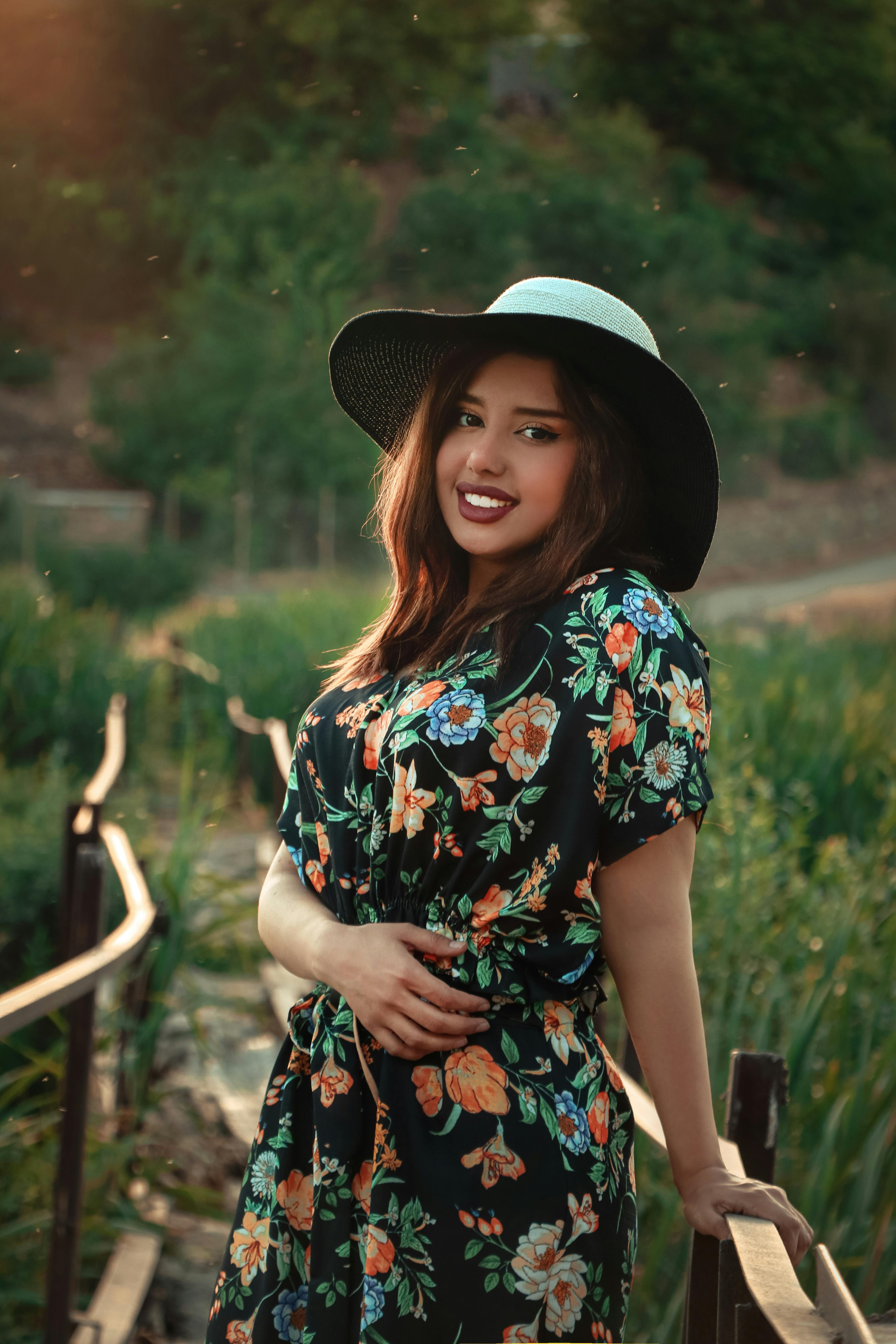 Young woman posing in floral dress and hat, smiling in a scenic outdoor setting.