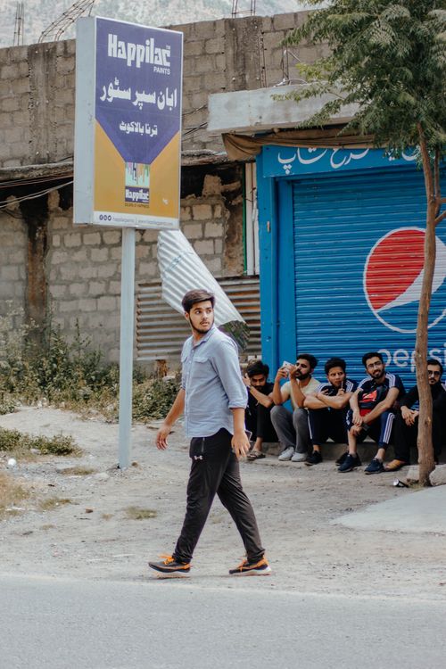 Free A young man walks down a street with a group sitting in the background. Urban setting with a billboard and vibrant colors. Stock Photo