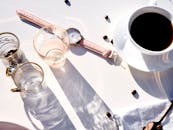 白色背景上，一杯咖啡、一只粉红手表和一个玻璃杯构成了时尚的咖啡场景