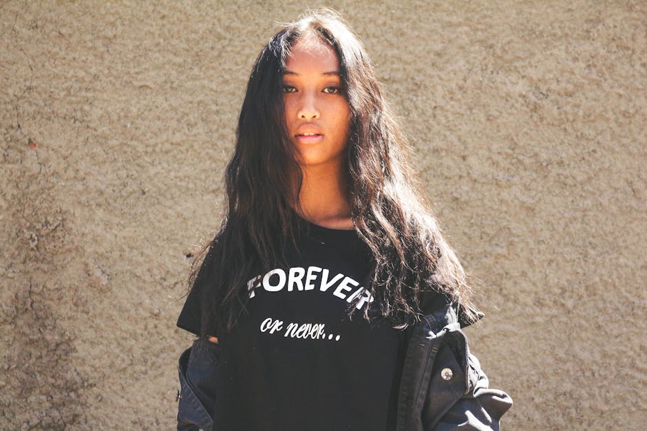 Portrait of a young woman with long hair posing against a textured wall, wearing a black t-shirt in sunlight.
