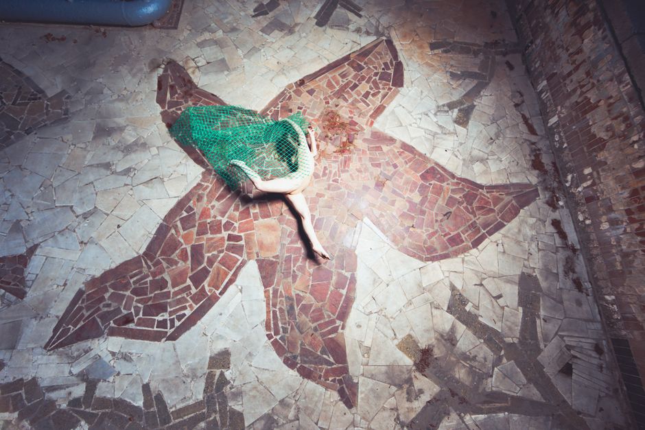 A woman wrapped in green netting lies on a geometric mosaic floor, creating an artistic visual.
