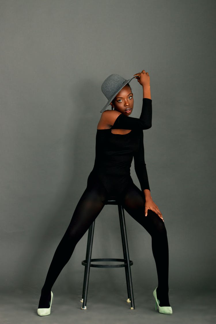 Woman Fashion Model In Black Tight Outfit And Hat Sitting On Stool And Posing