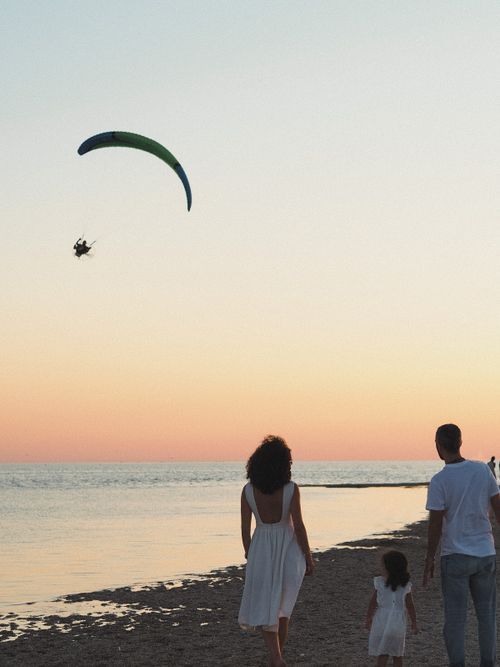 Free A family enjoying a peaceful walk on the beach at sunset with a paraglider in the sky. Stock Photo
