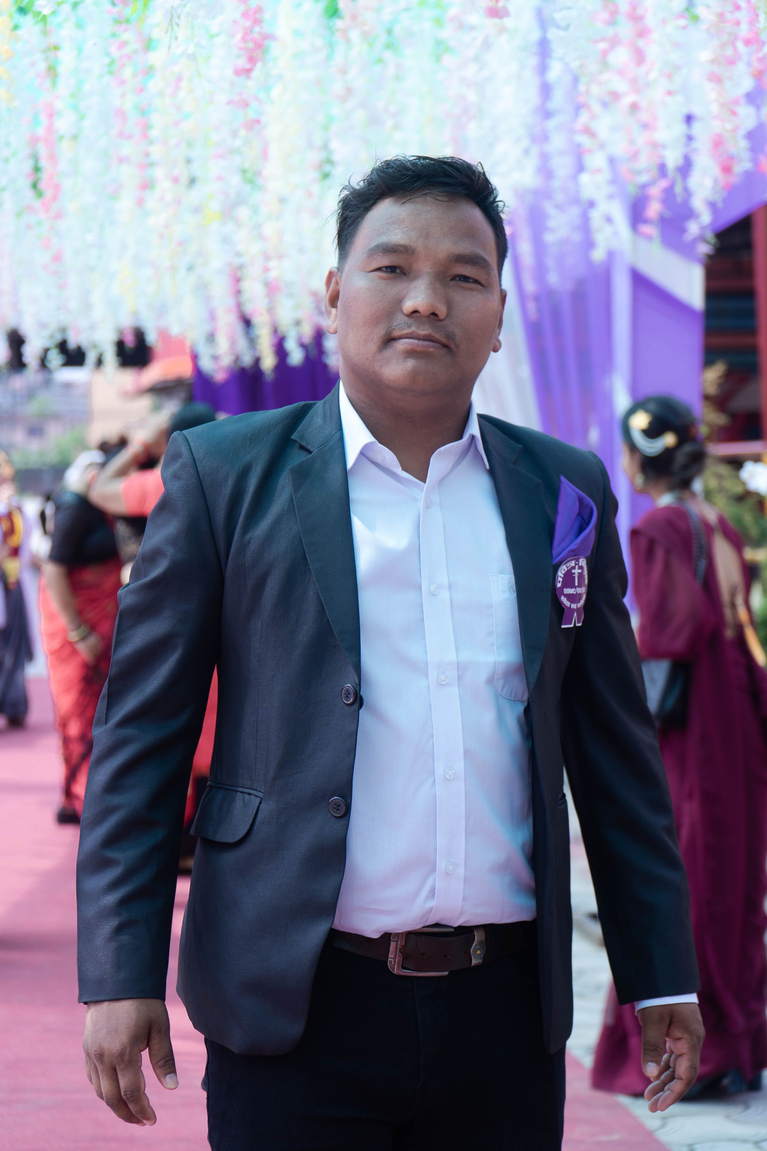 A confident Asian man in a black blazer and white shirt at a formal event with a colorful background.