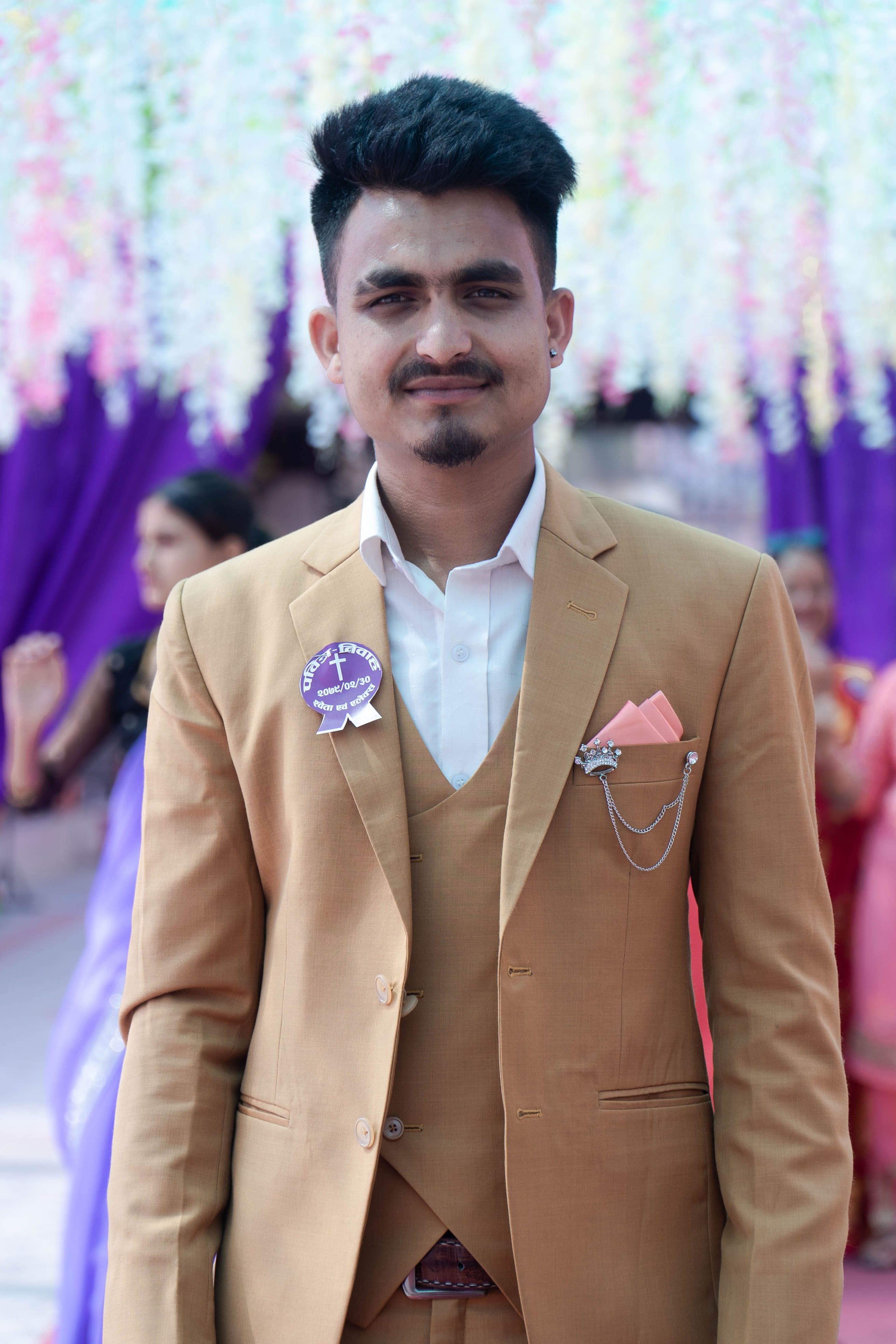 A young man in a tan suit with a boutonniere stands confidently at an outdoor event with colorful decorations.