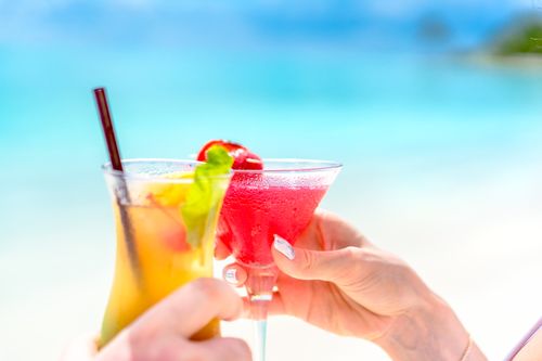 Free Two vibrant cocktails held by hands on a sunny beach with turquoise water. Stock Photo