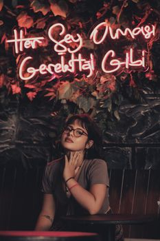 Stylish portrait of a young woman in glasses posing under neon lights in Fethiye, Turkey.