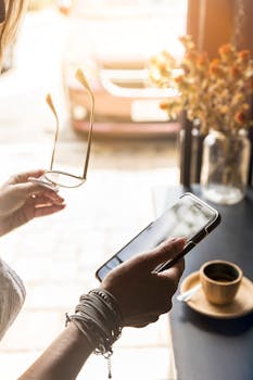 一名妇女在室内阳光直射下，拿着智能手机和眼镜，站在咖啡杯旁