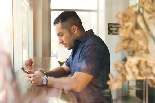 Selective Focus Photography of Man Sitting in Front of Brown Wooden Table While Holding Smartphone