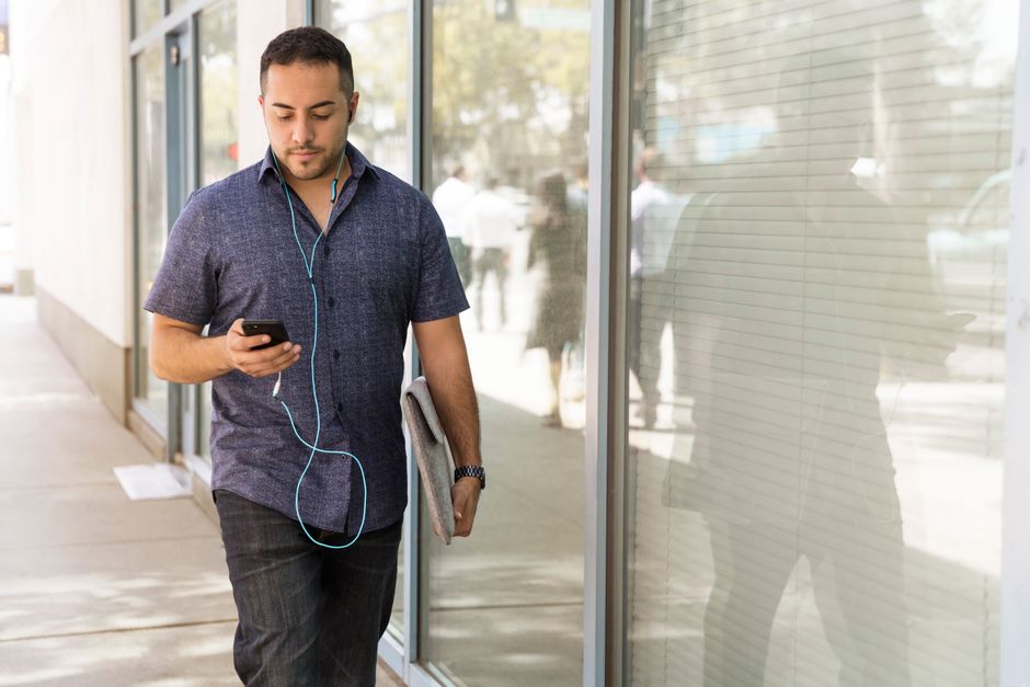 A young man with earphones walks along a glass-paneled urban street, holding a smartphone. A young man with earphones walks along a glass-paneled urban street, holding a smartphone.