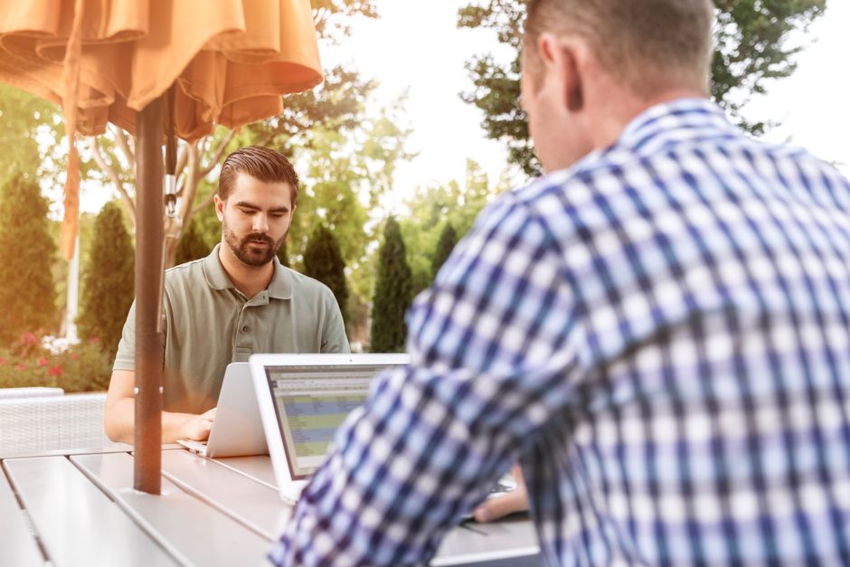 Men working on laptops outdoors, enjoying remote work and fresh air under a patio umbrella. Men working on laptops outdoors, enjoying remote work and fresh air under a patio umbrella.