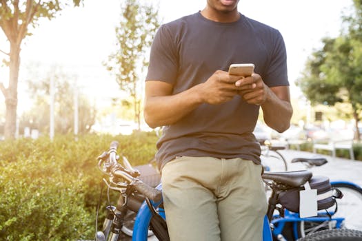 Casual young man leaning on a bike outdoors while using a smartphone in daylight.
