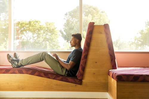 Free Man Sitting on Maroon and Brown Fabric Padded Sofa Using Laptop Beside Glass Window Stock Photo