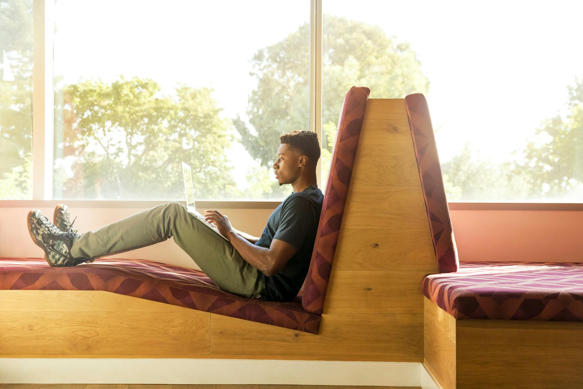 A young man lounging on a sofa, working remotely from his laptop in a bright room.