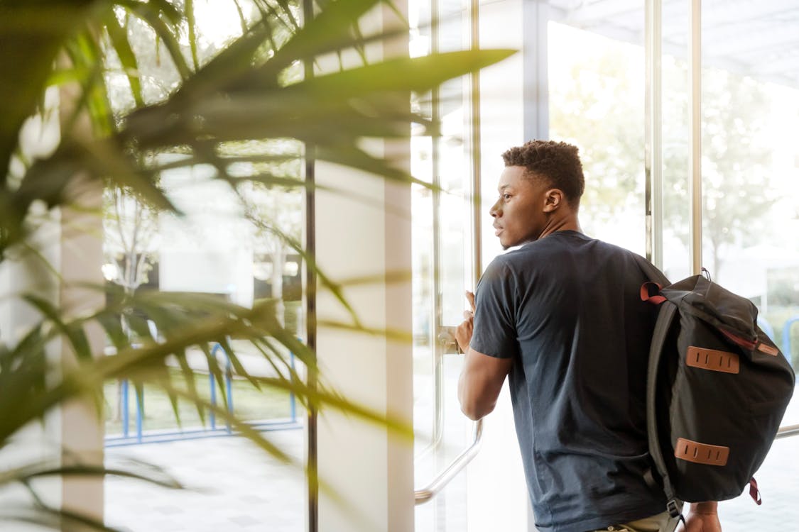 Free Man Looking Outside Window Carrying Black and Brown Backpack While Holding His Hand on Window Stock Photo