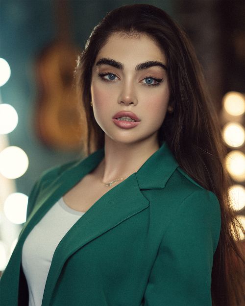 Free Beautiful woman in a green blazer posing confidently with soft bokeh lights behind her. Stock Photo