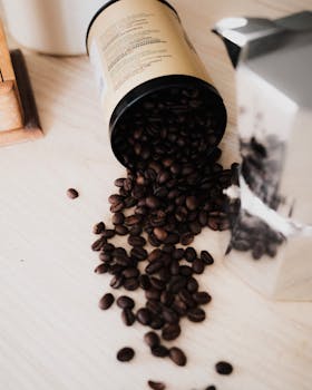 Coffee beans spilling from a container onto a wooden surface beside a metallic coffee maker.