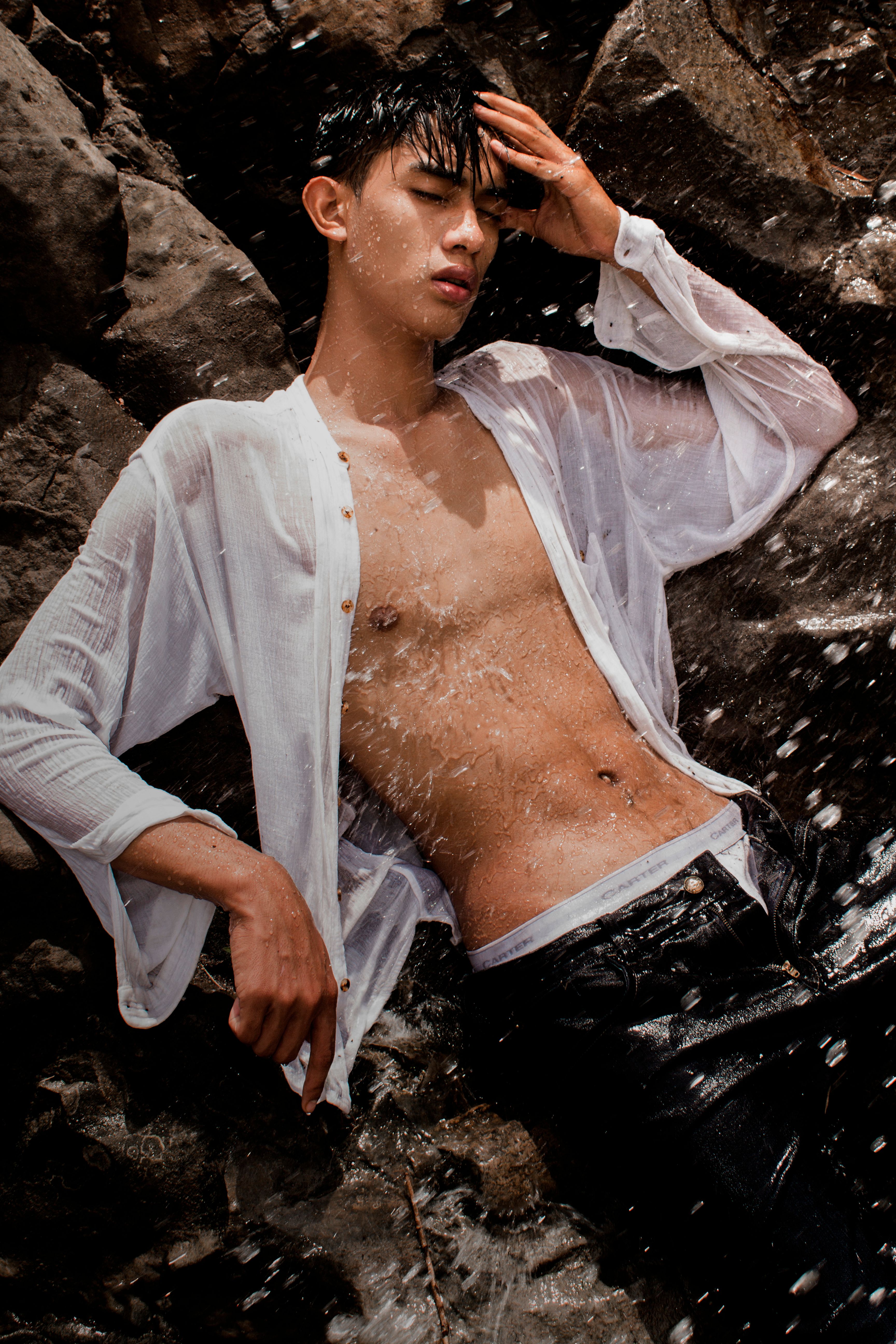 Shirtless Asian man in white sleeves posing confidently by a waterfall.