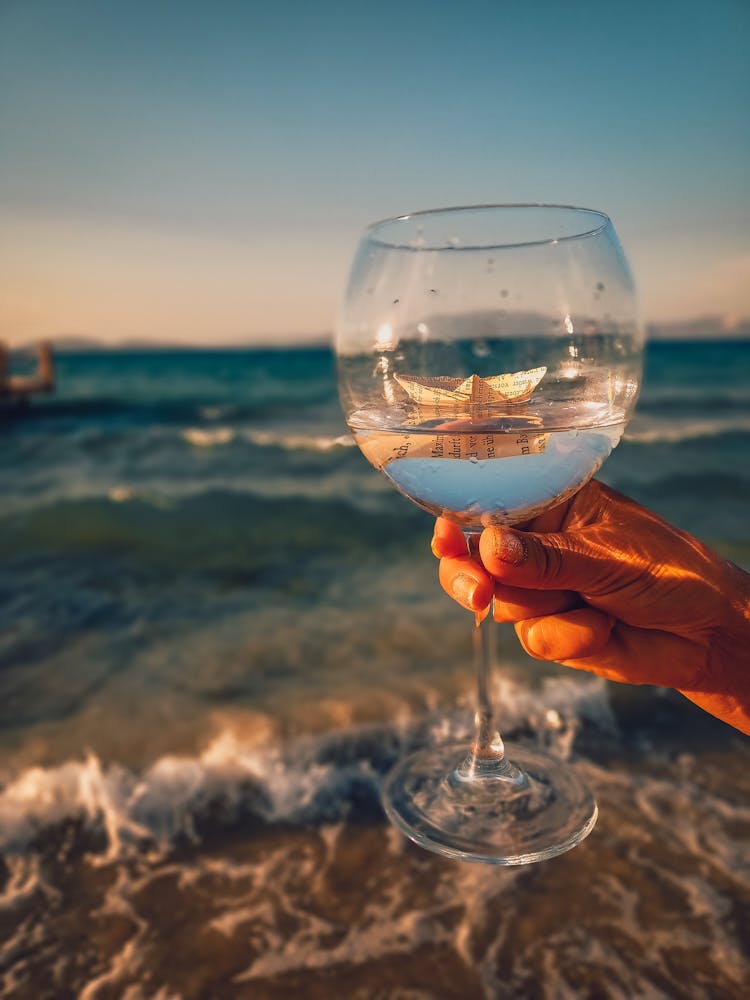 Person Holding Clear Wine Glass With Paper Boat And Water Inside 