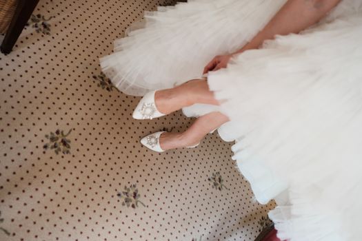 A close-up of a bride in white heels and gown on patterned carpet, ready for a special day.