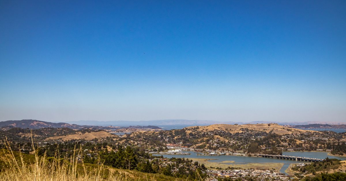 A picturesque landscape view of a town with rolling hills and a river under a clear blue sky.