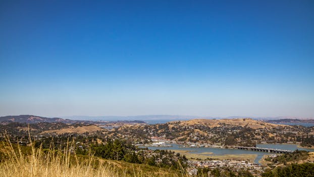 A picturesque landscape view of a town with rolling hills and a river under a clear blue sky.