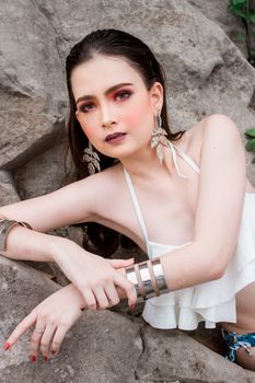 Elegant woman in white bikini poses against rocky backdrop, showcasing statement jewelry.