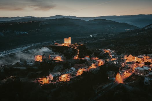 A breathtaking aerial shot capturing an illuminated hilltop village and castle at twilight surrounded by mountains.