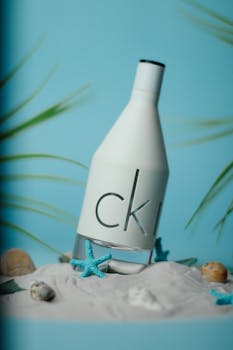 Perfume bottle on white sand with seashells and starfish creating a minimalist coastal scene.