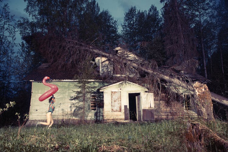 Woman With Inflatable Flamingo Pool Float Running By An Abandoned House With Fallen Tree