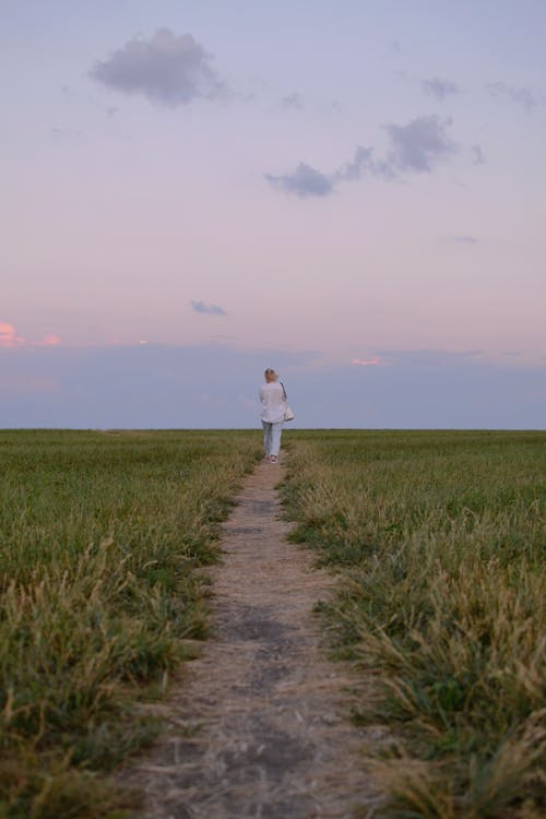 Person in White Long Sleeves Walking on a Pathway In the Middle of Grass Field 