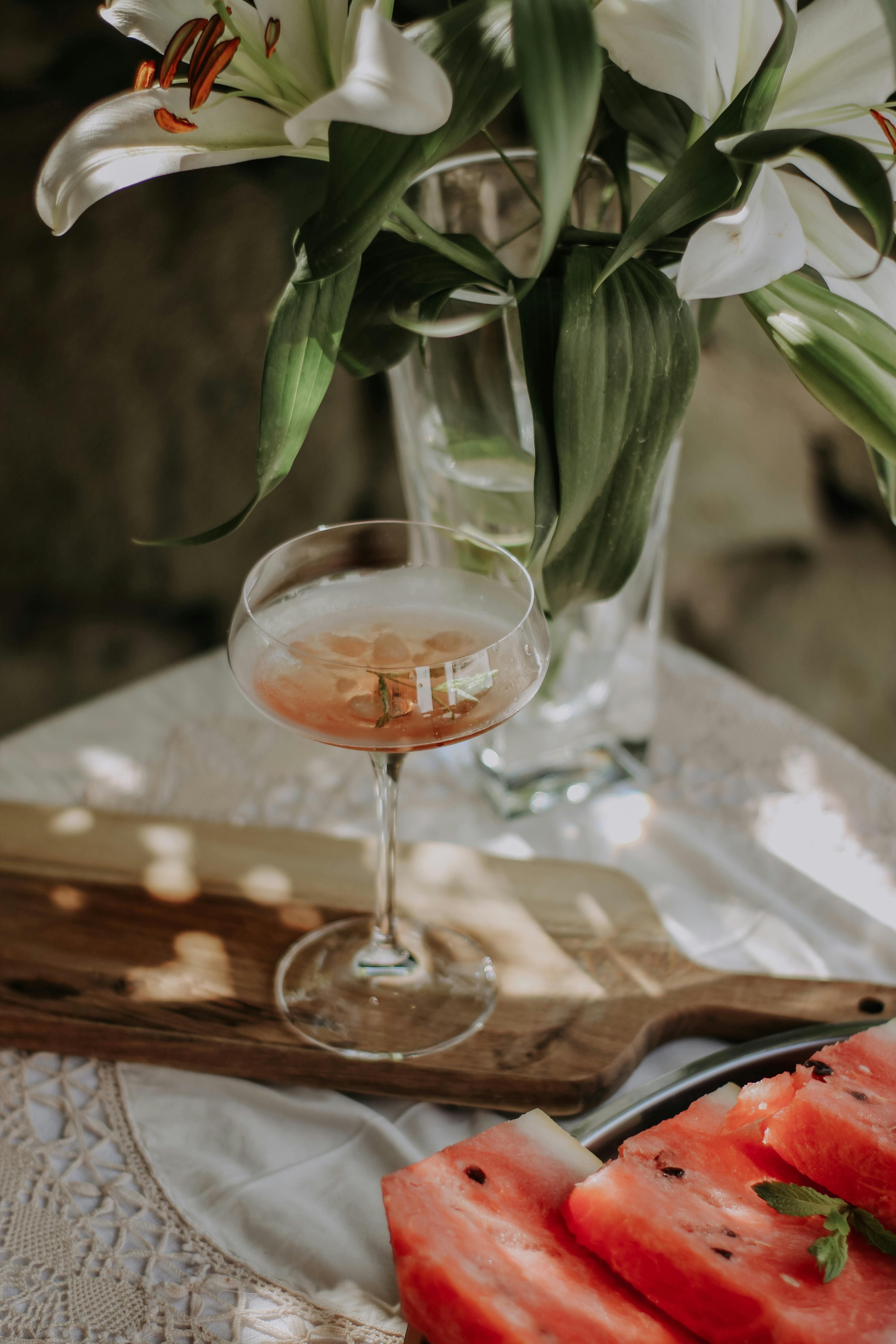 Elegant cocktail with watermelon slices and lilies on a sunlit table.