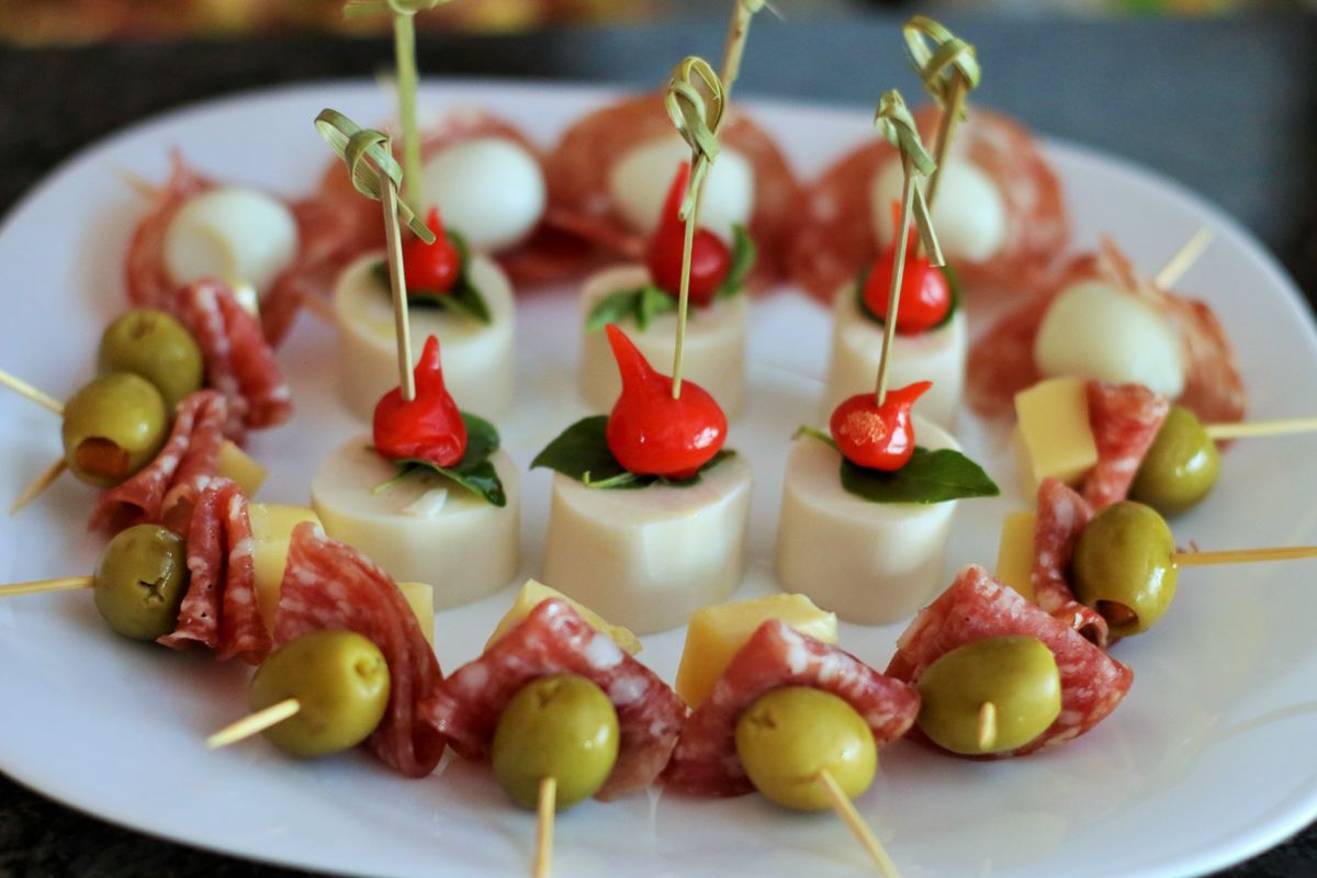 White platter of cocktail skewers with salami, green olives, cheese cubes, mozzarella and red peppers