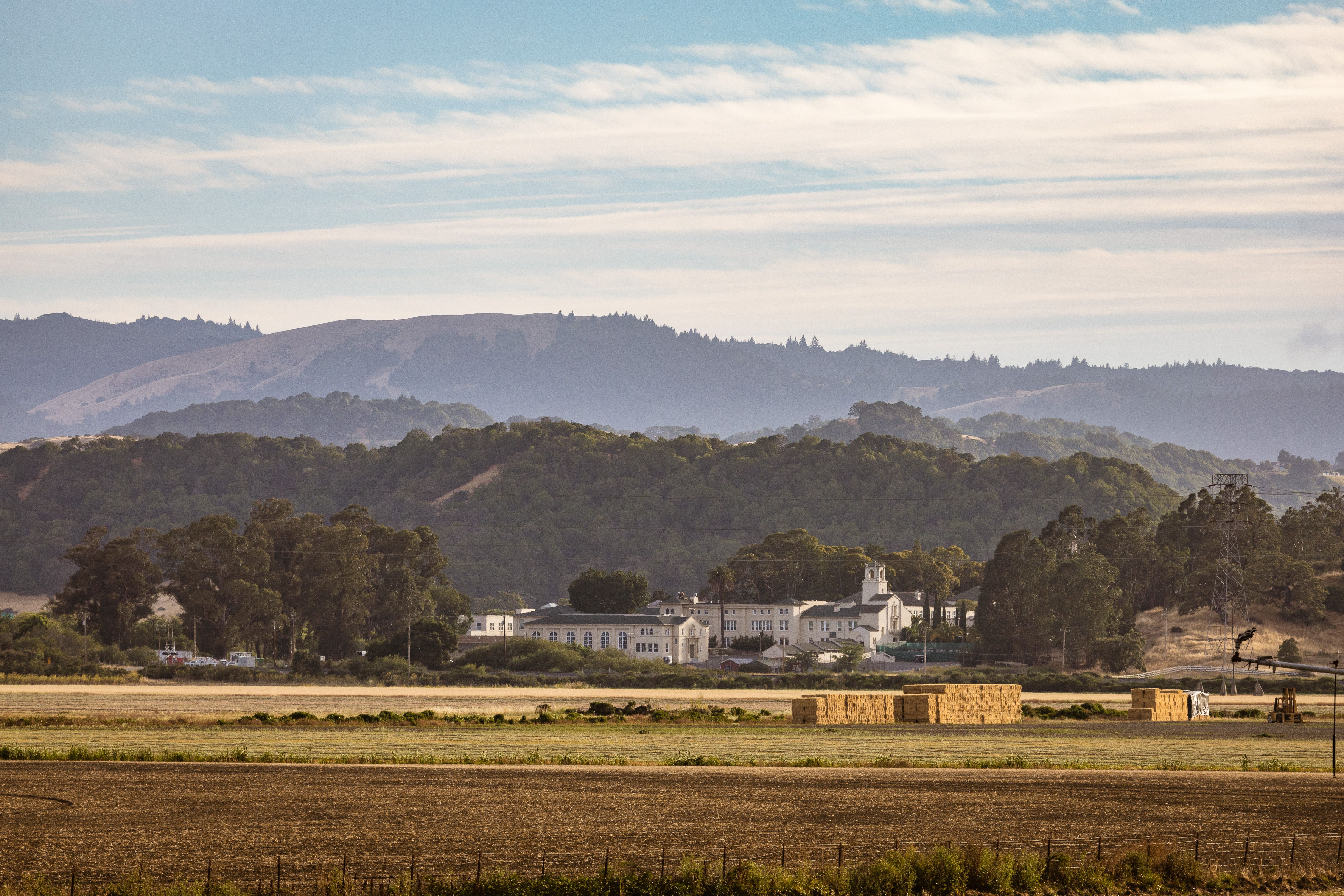 Tranquil rural scene featuring haystacks, farmland, and distant hills under a blue sky.