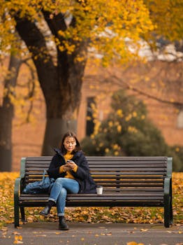 A woman uses her phone while sitting on a bench surrounded by autumn leaves in a city park.